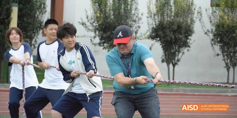 A man in a green shirt and a black baseball cap pulling a thick rope during a tug-of-war event with students. Beijing No.4 High School International Courses Jialian Campus.