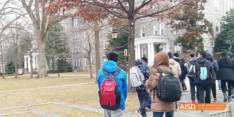 A group of students, including one with a bright blue backpack, walking past historic stone buildings on campus. Beijing No.4 High School International Courses Jialian Campus.