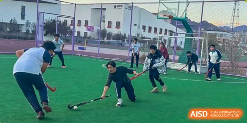 Students playing a game that looks like field hockey or a similar ground sport on a green pitch. Beijing No.4 High School International Courses Jialian Campus.