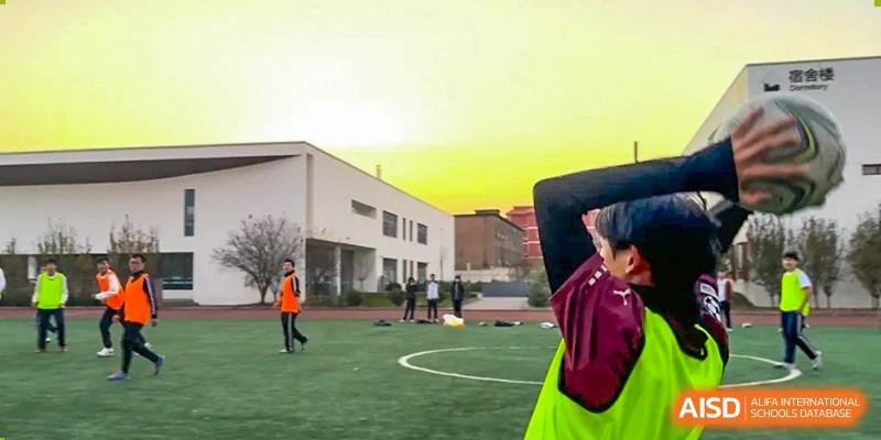A student running on an outdoor track next to a green field with a white building in the background at sunset. Beijing No.4 High School International Courses Jialian Campus.