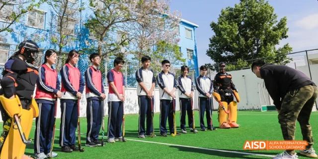 A group of students in white and dark blue uniforms lined up on a green field next to a light blue school building. Beijing No.4 High School International Courses Jialian Campus.
