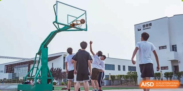 Students playing basketball on an outdoor court, with one student reaching up to shoot toward the hoop. Beijing No.4 High School International Courses Jialian Campus.