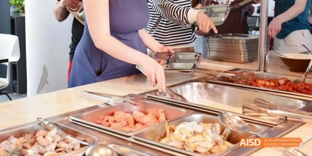 A close-up shot of a person serving themselves food from a buffet line in a cafeteria. Beijing No.4 High School International Courses Jialian Campus.