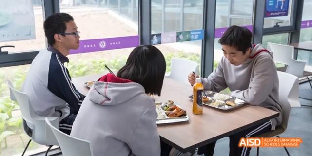 A student with a red and white jacket seated at a table, eating a meal and drinking from a bottle. Beijing No.4 High School International Courses Jialian Campus.
