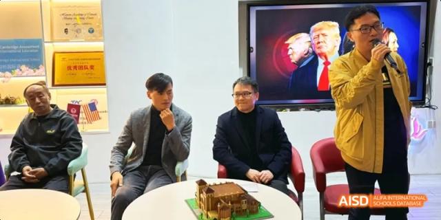 Two men sitting and talking near a small cake, with a digital screen showing a picture in the background. Beijing No.4 High School International Courses Jialian Campus.
