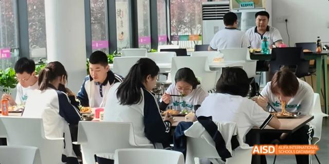 Students in uniform seated at modern white tables, eating together in a brightly lit dining area. Beijing No.4 High School International Courses Jialian Campus.