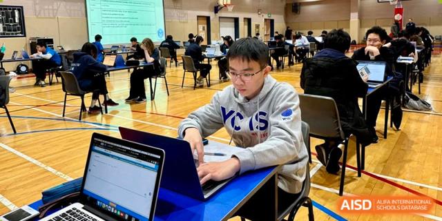 A male student wearing a school hoodie working on a laptop at a desk in a classroom at Yew Chung International School of Beijing.