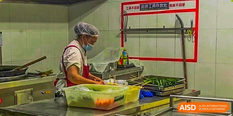 A kitchen staff member wearing a mask and hairnet preparing food, likely in the school cafeteria or kitchen at Maple Leaf International School Chongqing.