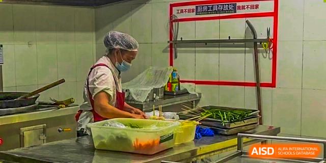 A kitchen staff member wearing a mask and hairnet preparing food, likely in the school cafeteria or kitchen at Maple Leaf International School Chongqing.