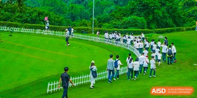 A group of people on a large, well-maintained green sports field, possibly practicing golf or other outdoor activities, at Maple Leaf International School Chongqing.