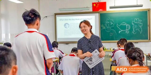 A smiling female teacher in a polka-dot dress addressing students in a bright, interactive classroom at Wuxi Big Bridge Academy.