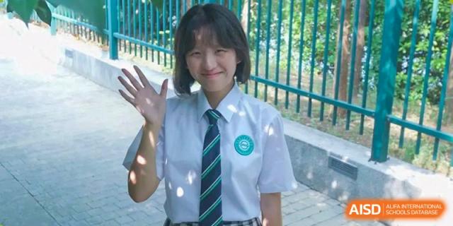 A smiling female student in her school uniform, waving outdoors at Wuxi Big Bridge Academy.