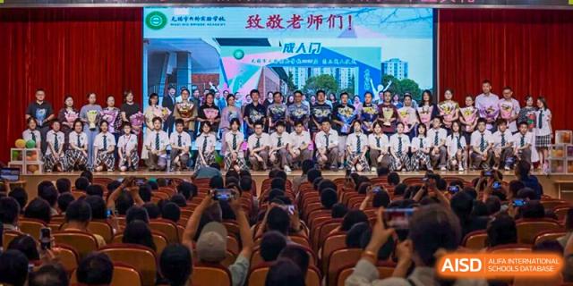A large group of students in an auditorium cheering and waving to teachers, possibly for a Teacher Appreciation Day event at Wuxi Big Bridge Academy.