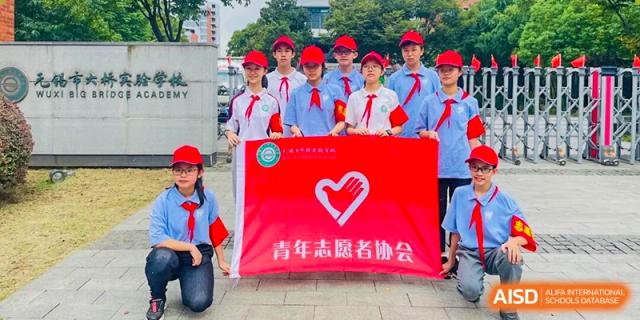 A group of students in red scarves holding a banner for the Youth Volunteer Association at Wuxi Big Bridge Academy.