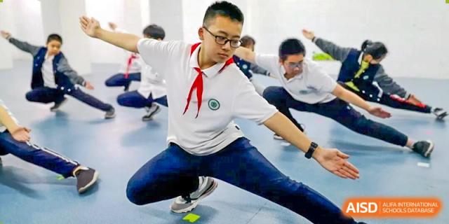 A male student wearing a red scarf performing a dynamic stretch or martial arts move during a physical education class at Wuxi Big Bridge Academy.
