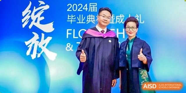 The principal of Zhuhai Oriental Yinghua International Academy posing in a graduation gown in front of a 2024 commencement banner.