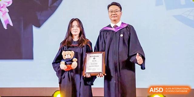 A graduate from Zhuhai Oriental Yinghua International Academy and a teacher posing together, holding an award and a graduation teddy bear.