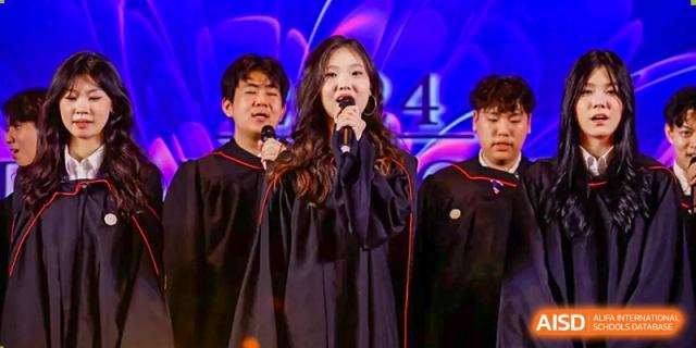 A female graduate from Zhuhai Oriental Yinghua International Academy singing into a microphone on stage during a ceremony, flanked by other graduates.