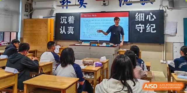 A male teacher points to a screen with Chinese text while addressing a class of seated students in a bright classroom at Changjun Bilingual School Of Changsha City.