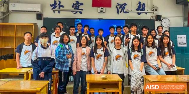 A group of students are posing and smiling for a photo together at the front of a classroom, standing in front of a large screen and traditional Chinese wall script at Changjun Bilingual School Of Changsha City.