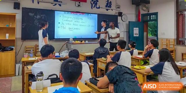 An international teacher (male) and a student interact during a lesson, writing on an illuminated whiteboard in a classroom setting with several students visible from the back at Changjun Bilingual School Of Changsha City.