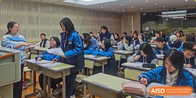 A female student standing in the aisle of a classroom, appearing to speak or instruct a large group of seated classmates at Changjun Bilingual School Of Changsha City.