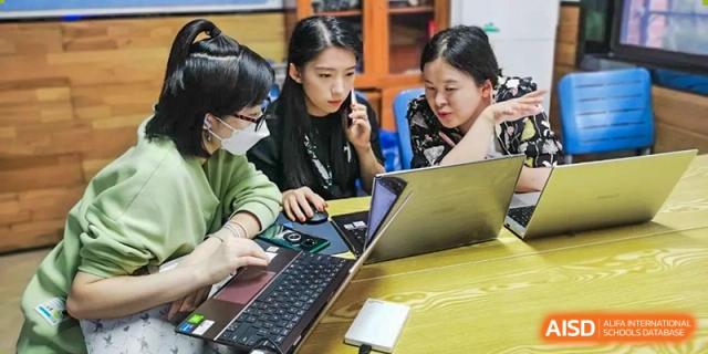 A small group of students huddle around a table, actively working on a laptop and a smartphone, collaborating on a project at Changjun Bilingual School Of Changsha City.