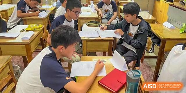 Several male students are deeply focused on studying at wooden desks, writing notes and reviewing papers in a classroom at Changjun Bilingual School Of Changsha City.