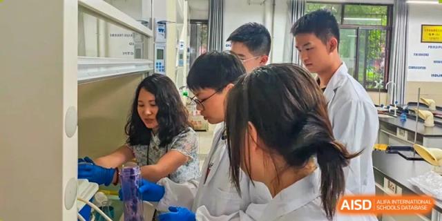 A group of students wearing safety goggles and white lab coats are clustered around a bench, performing a chemistry experiment with beakers and colored liquids at Changjun Bilingual School Of Changsha City.