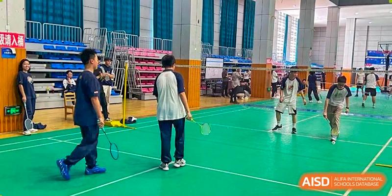 Two students from Changjun Bilingual School Of Changsha City standing on an indoor green badminton court, with one student viewed from the back, holding a racket.
