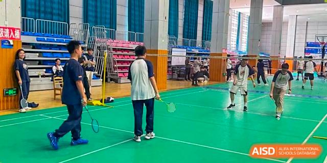 Two students from Changjun Bilingual School Of Changsha City standing on an indoor green badminton court, with one student viewed from the back, holding a racket.