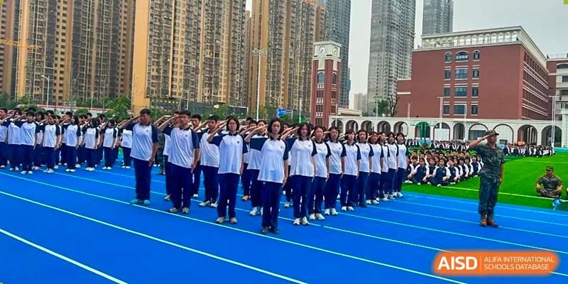 A large group of students from Changjun Bilingual School Of Changsha City in uniform, lined up in a salute or formation on a blue track outdoors with tall buildings in the background.