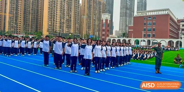 A large group of students from Changjun Bilingual School Of Changsha City in uniform, lined up in a salute or formation on a blue track outdoors with tall buildings in the background.