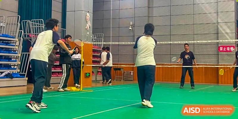 A student from Changjun Bilingual School Of Changsha City playing badminton on a green court indoors, viewed from the back during a rally.