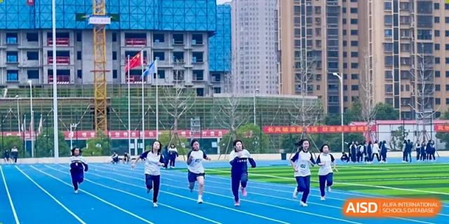 Students in sportswear are running on a blue track on a sports field at Changjun Bilingual School Of Changsha City, with school and residential buildings in the background.