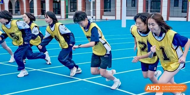 Two students from Changjun Bilingual School Of Changsha City running on a blue track, one wearing a yellow bib, in a close-up action shot.