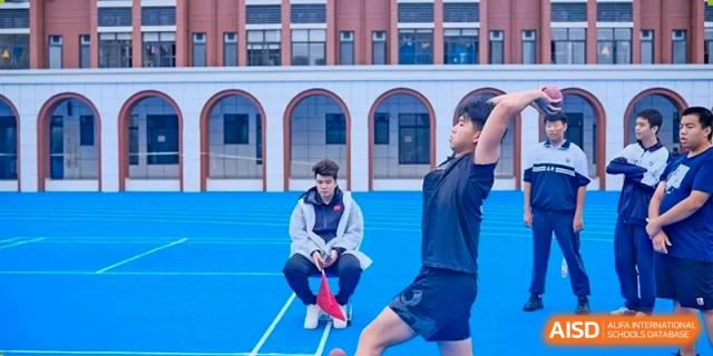 Two male students are on a blue running track, one sitting and one standing, potentially playing or practicing a sport, outside the academic buildings of Changjun Bilingual School Of Changsha City.