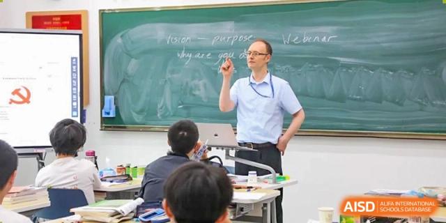 A teacher from Cogdel Cranleigh High School lecturing in a classroom, standing in front of a traditional blackboard with writing.