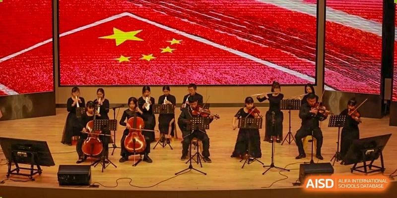 A small student orchestra from Cogdel Cranleigh High School playing string instruments on a stage beneath a banner featuring the Chinese flag.