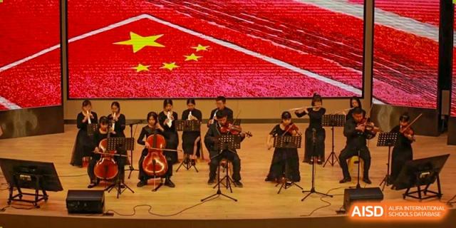 A small student orchestra from Cogdel Cranleigh High School playing string instruments on a stage beneath a banner featuring the Chinese flag.