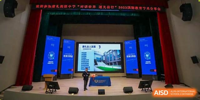 A teacher or speaker standing on a stage next to a large presentation screen during an event at Cogdel Cranleigh High School.