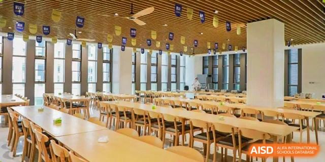 A bright and spacious dining hall or cafeteria at Cogdel Cranleigh High School, featuring wooden tables and chairs and decorated with banners.