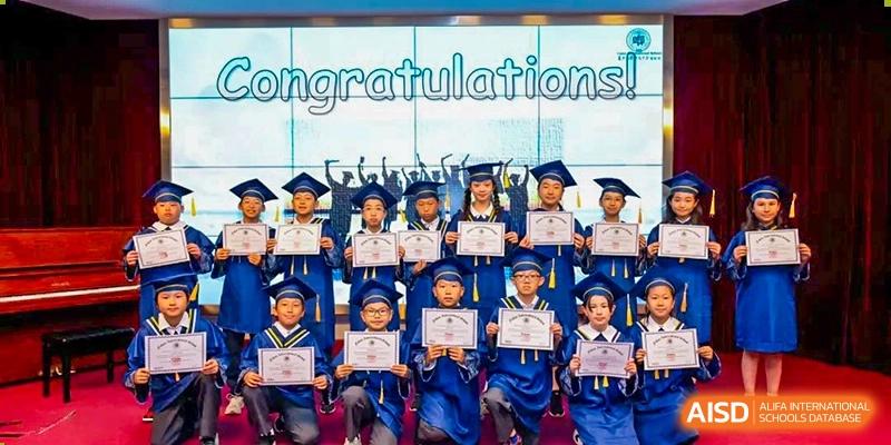 A group of happy Fudan International School graduates in caps and gowns holding up their diplomas beneath a "Congratulations" banner.
