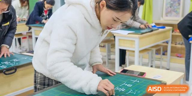A female teacher from Humen Foreign Language School International Division in a white fluffy sweater works at a desk, carefully measuring or drawing on a piece of paper.