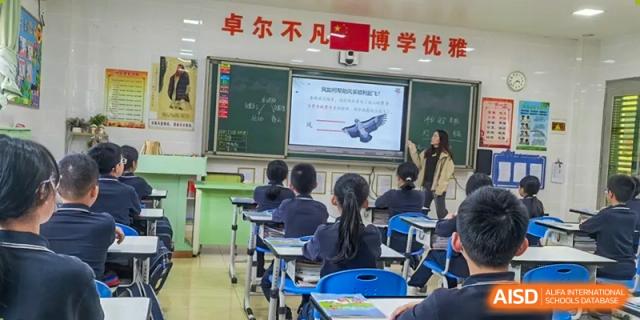 A teacher from Humen Foreign Language School International Division stands next to a smart board displaying a lesson to students seated in a classroom.