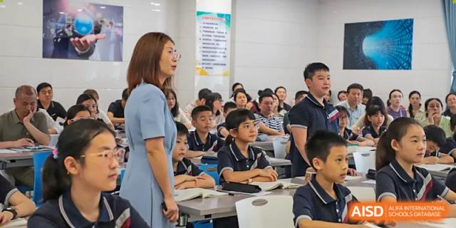 A female teacher from Humen Foreign Language School International Division stands beside her desk, teaching a class of students.