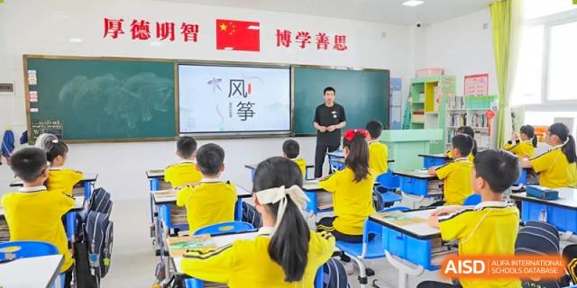 A teacher from Humen Foreign Language School International Division stands at the front of a classroom with a smart board, teaching a lesson to students wearing yellow shirts.