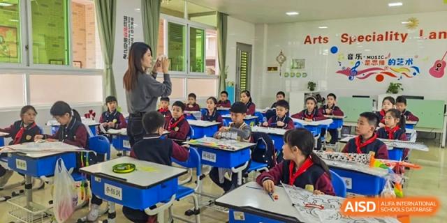Students from Humen Foreign Language School International Division sit in a brightly lit classroom with a teacher standing near the front and an "Arts" sign on the wall.