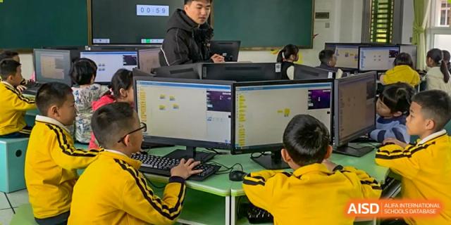 Students from Humen Foreign Language School International Division in yellow shirts work on computers with multiple screens in a school computer lab.
