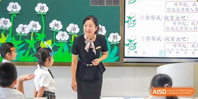 A female teacher from Humen Foreign Language School International Division wearing a black uniform and a patterned scarf stands in front of a green chalkboard, smiling at the class.